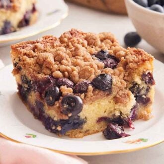 A close up view of a plate of blueberry buckle with fresh blueberries scattered in the front and a bowl of them in the background.