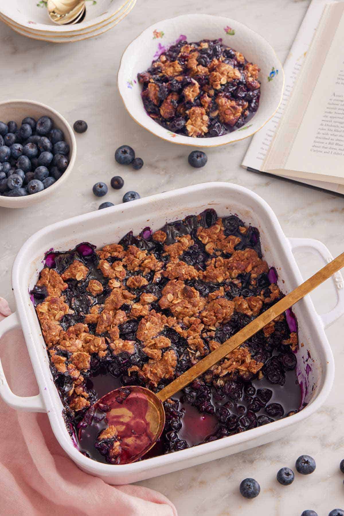 Overhead view of a white baking dish with blueberry crisp with a third scooped out. A serving in the bowl in the back along with a bowl of blueberries and a book.