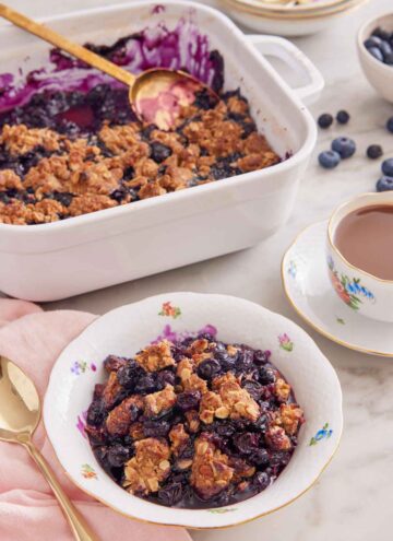 A bowl of blueberry crisp with a casserole dish with the rest of the dessert in the background also with a mug of tea.