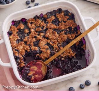 Pinterest graphic of an overhead view of a white baking dish filled with blueberry crisp with a third scooped out.