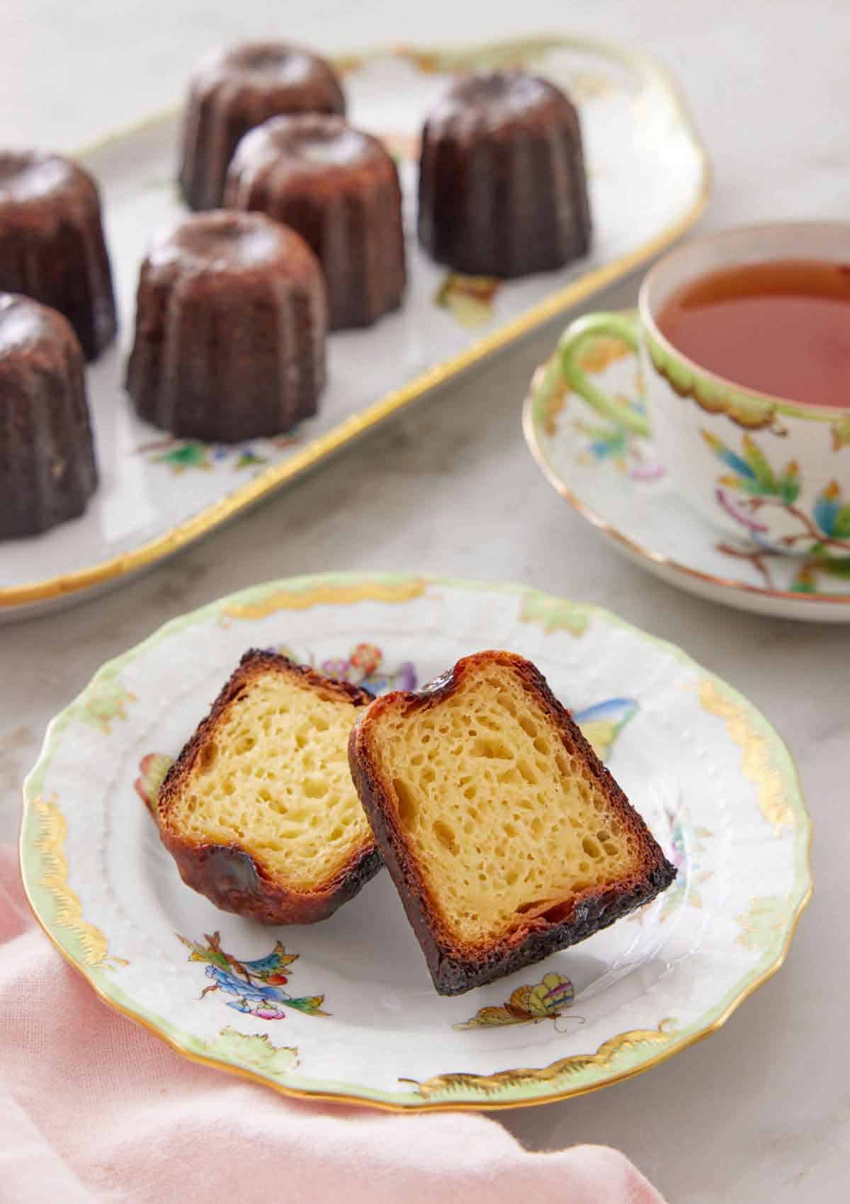 A plate with a canelé cut in half, showing the inside texture. Tea and a platte of canelés in the background.