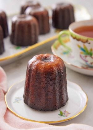 A canelé on a plate with more in a platter in the back along with a cup of tea.