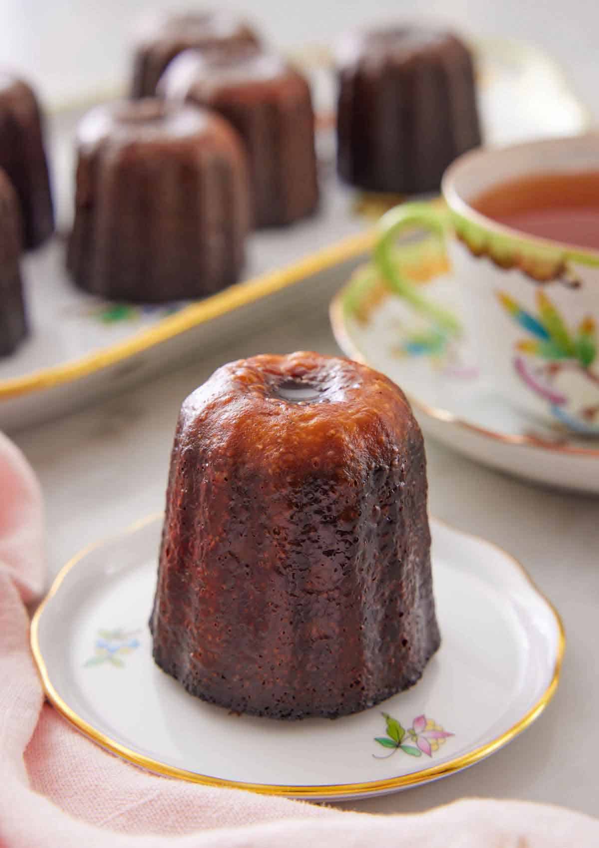 A canelé on a plate with more in a platter in the back along with a cup of tea.