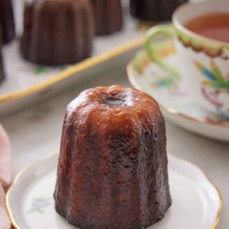 Pinterest graphic of a canelé on a plate with a cup of tea and more canelés on a platter in the background.
