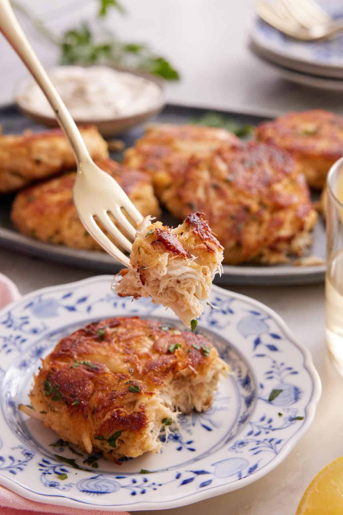 A fork lifting up a bite of crab cakes from the piece on the plate.