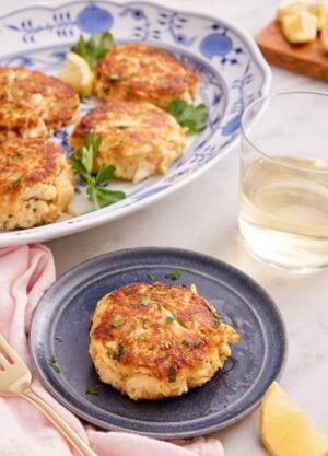 A small plate with a crab cake with a platter with more behind it and glass of wine.