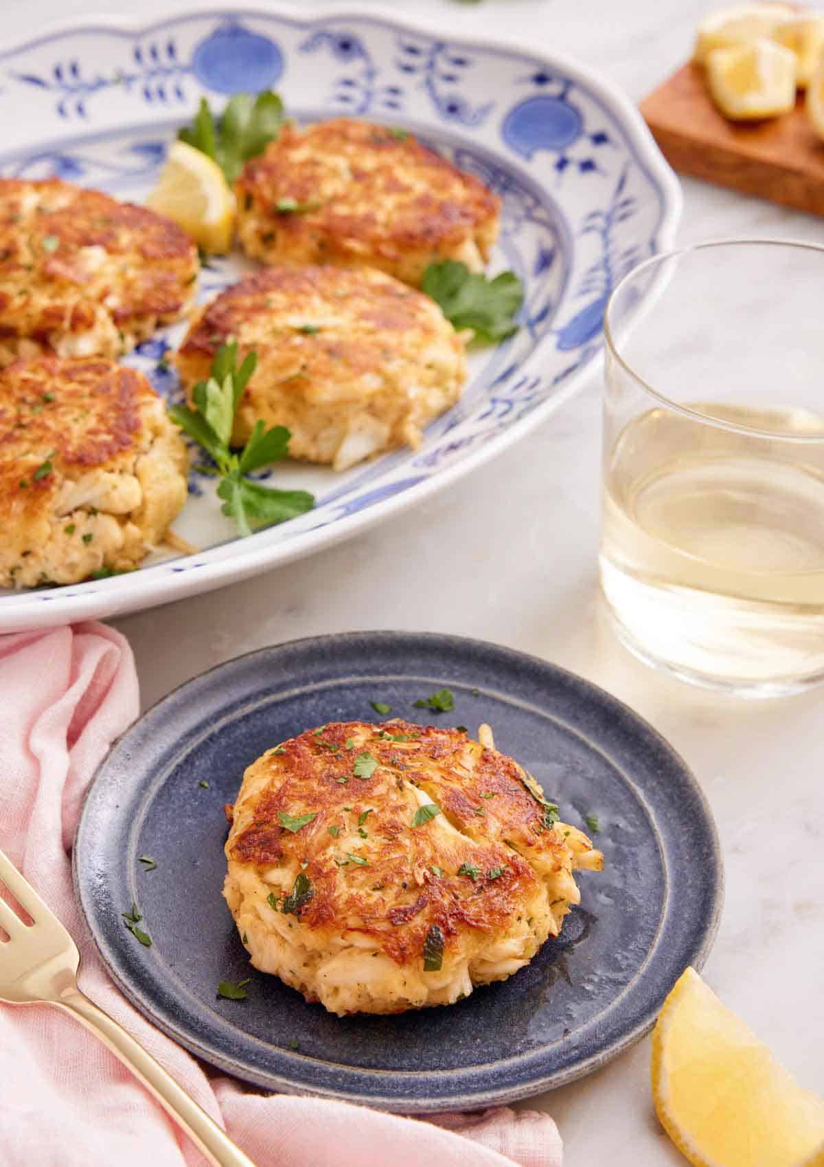 A small plate with a crab cake with a platter with more behind it and glass of wine.