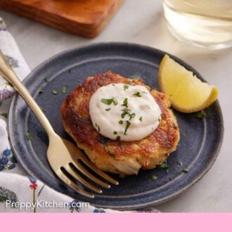Pinterest graphic of a plate with a crab cake topped with a dollop of tartar sauce with a lemon wedge and fork beside it.