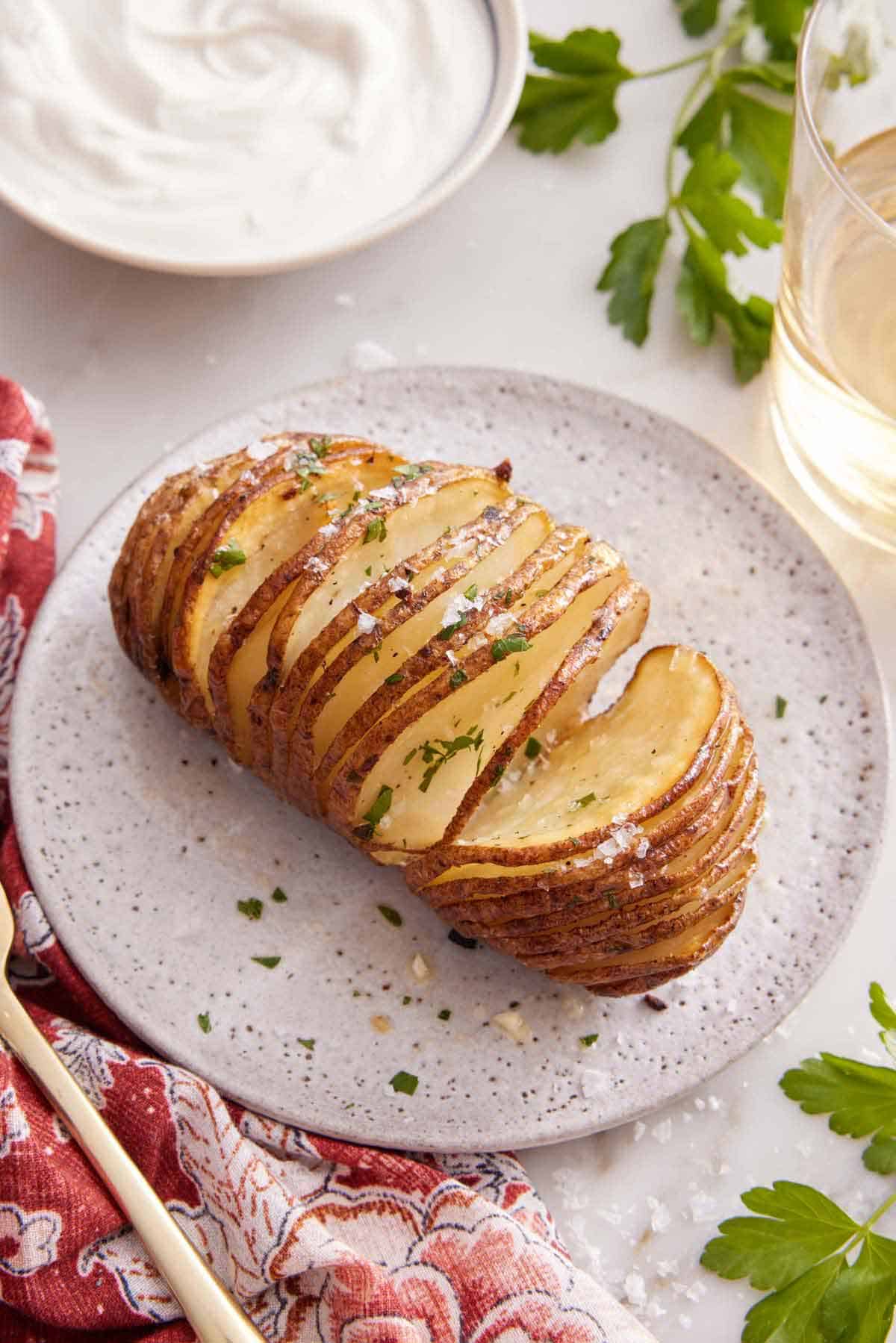 A white plate with a hasselback potato with a glass of wine and bowl of sour cream in the back.