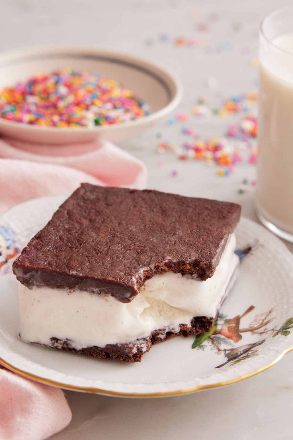 A plate with a ice cream sandwich with a bite taken out of the corner with a bowl of sprinkles in the background.