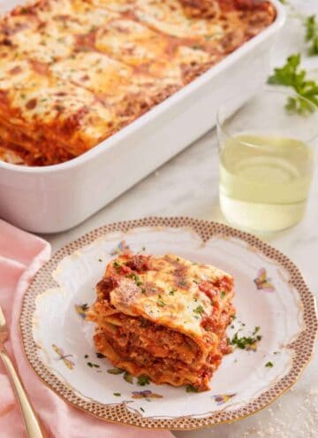 A slice of lasagna on a plate with a baking dish with the baked lasagna and a glass of wine in the background.