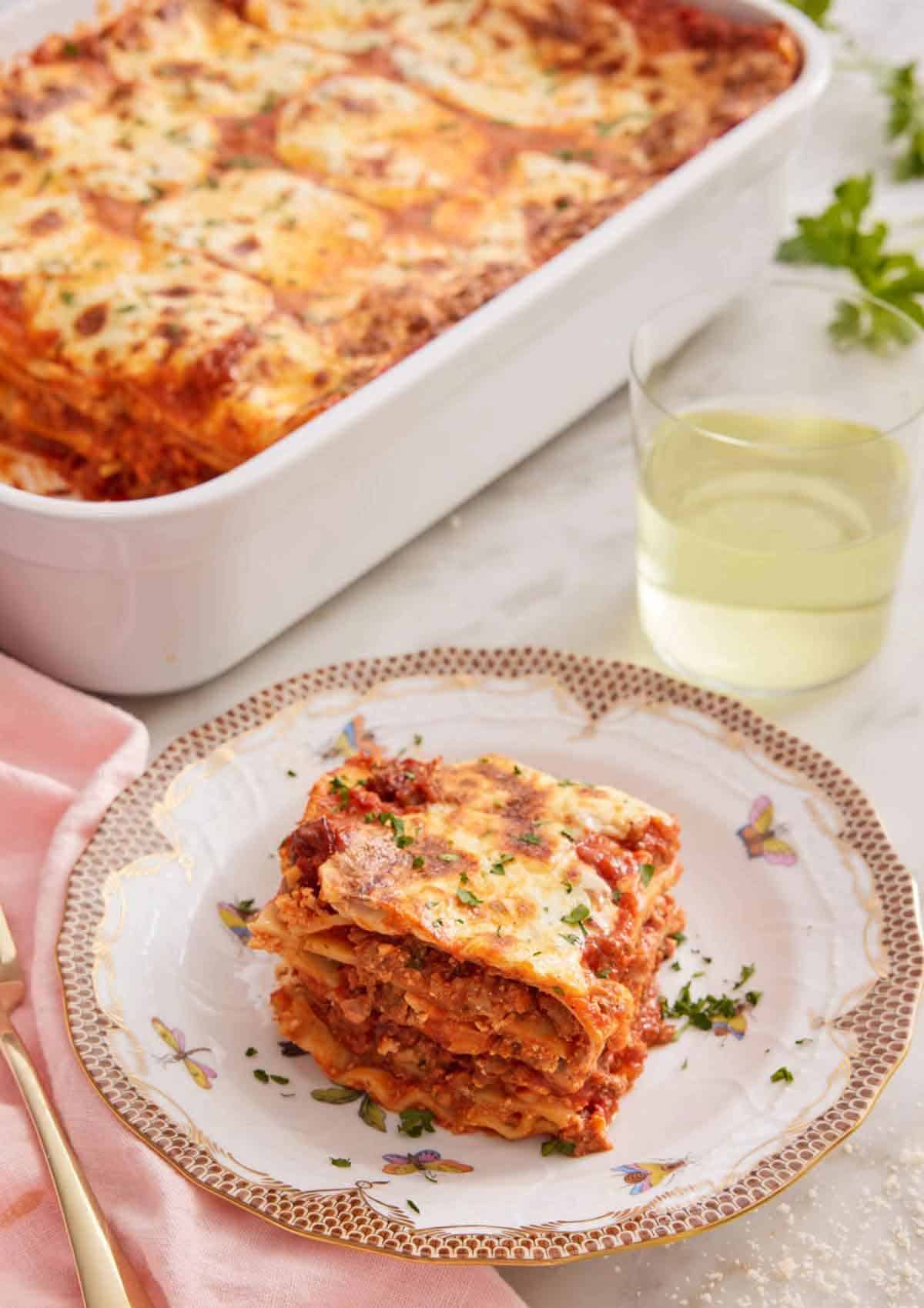 A slice of lasagna on a plate with a baking dish with the baked lasagna and a glass of wine in the background.