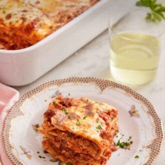 Pinterest graphic of a slice of lasagna on a plate with a glass of wine and a baking dish with more lasagna in the background.