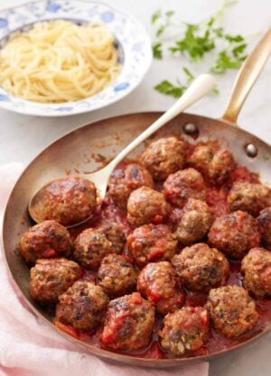 A skillet of meatballs with sauce and a spoon with some noodles in the background.