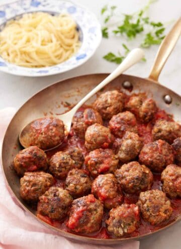 A skillet of meatballs with sauce and a spoon with some noodles in the background.