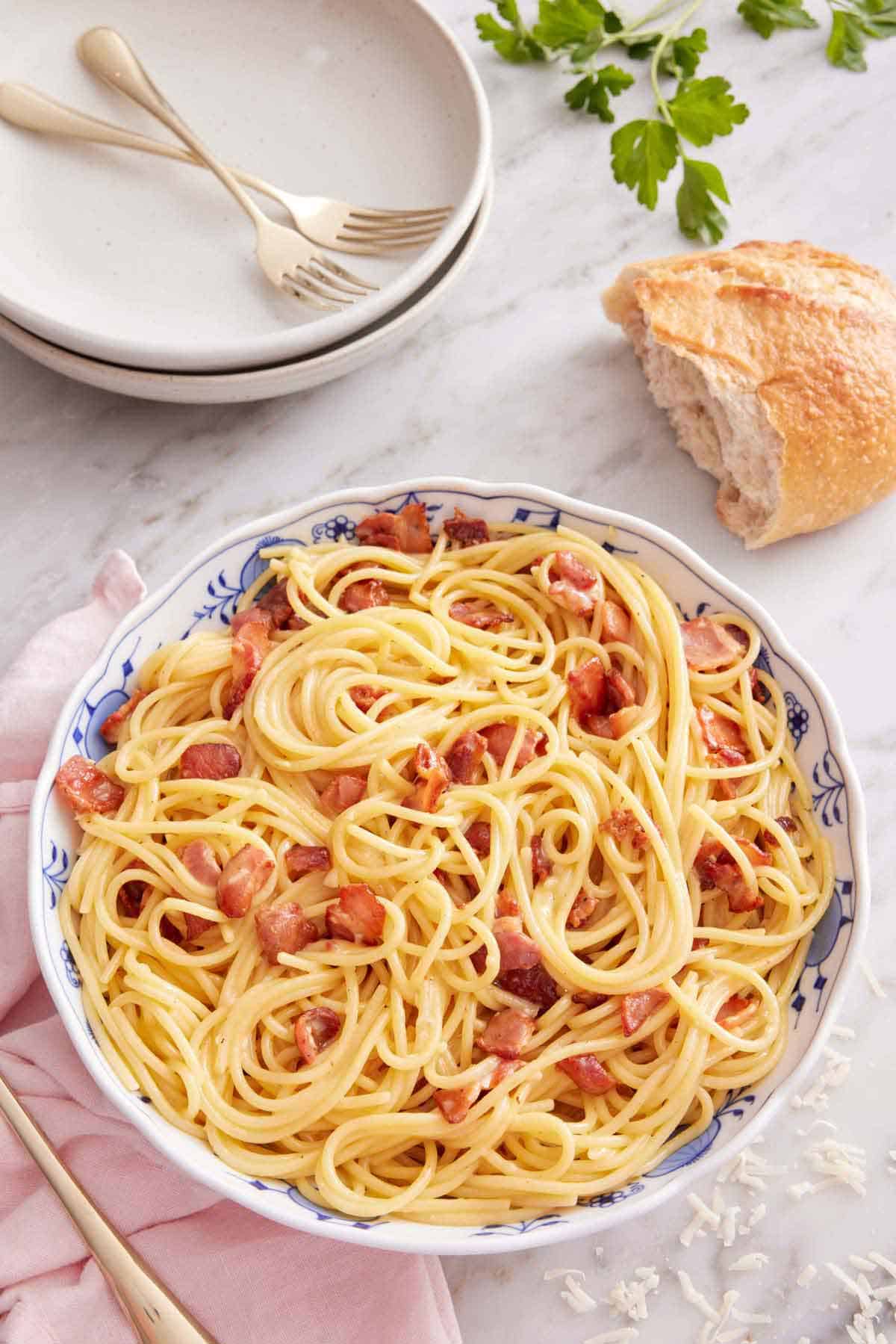 A bowl of pasta carbonara with torn bread and stacked plates with forks in the background.