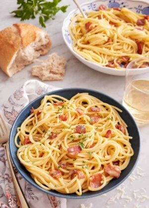 A bowl of pasta carbonara with torn bread, glass of wine, and another bowl of pasta in the back.