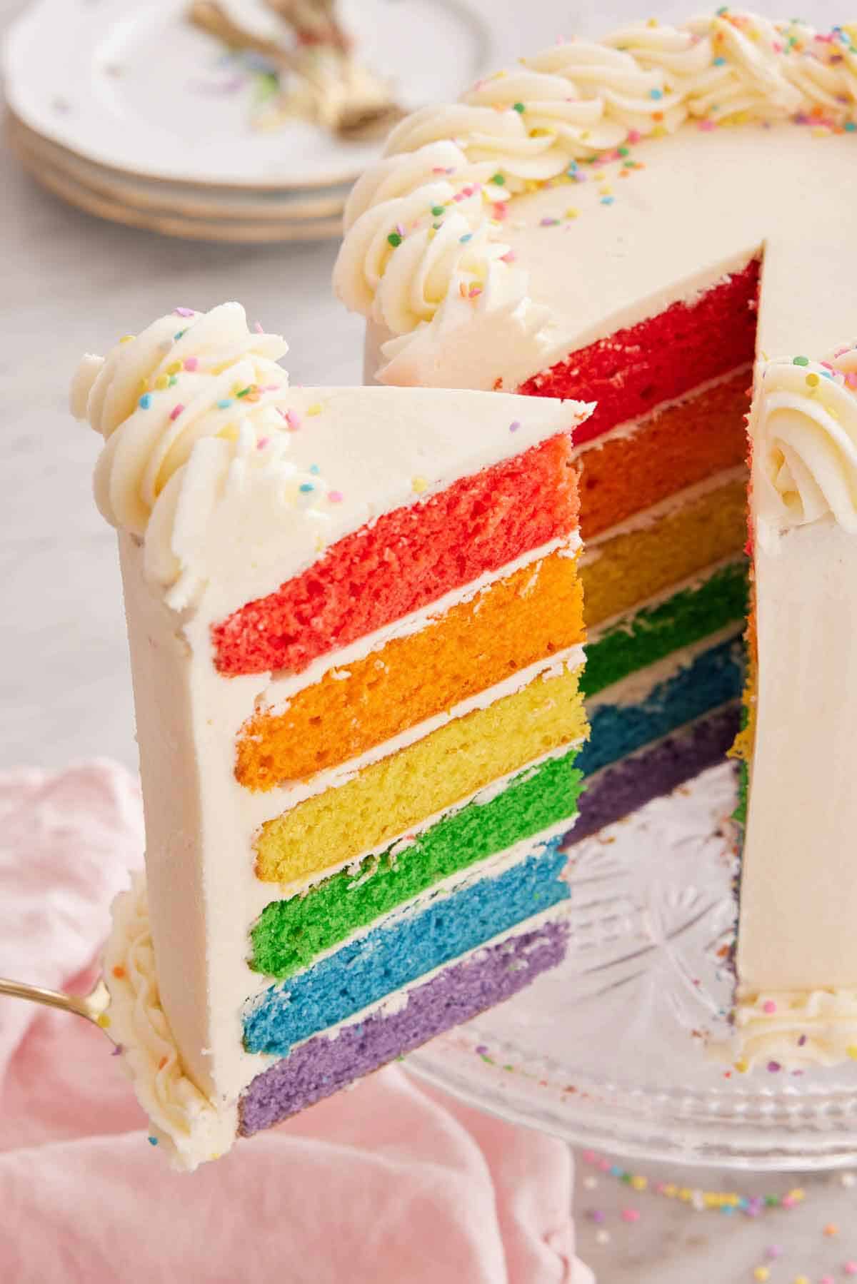 A slice of rainbow cake pulled with a cake spatula from the cake on the cake stand.