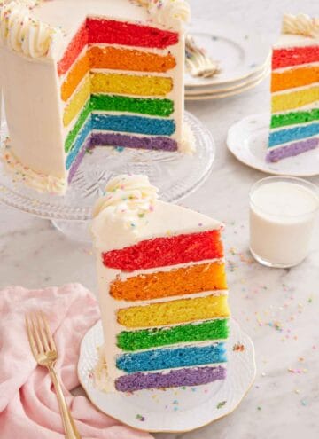 A plate with a slice of rainbow cake with a glass of milk, a second plated slice, and the cut cake on a cake stand in the background.
