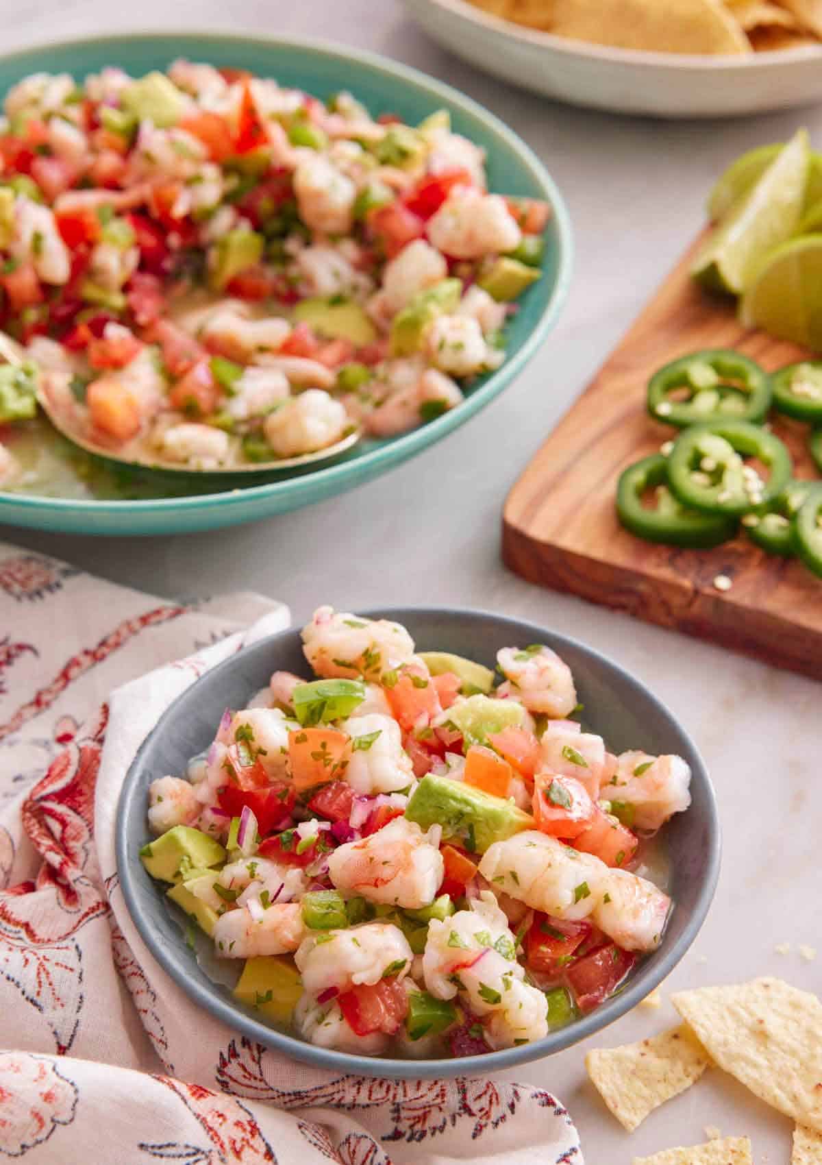 A bowl of shrimp ceviche with a platter in the back and cut garnishes on the side.