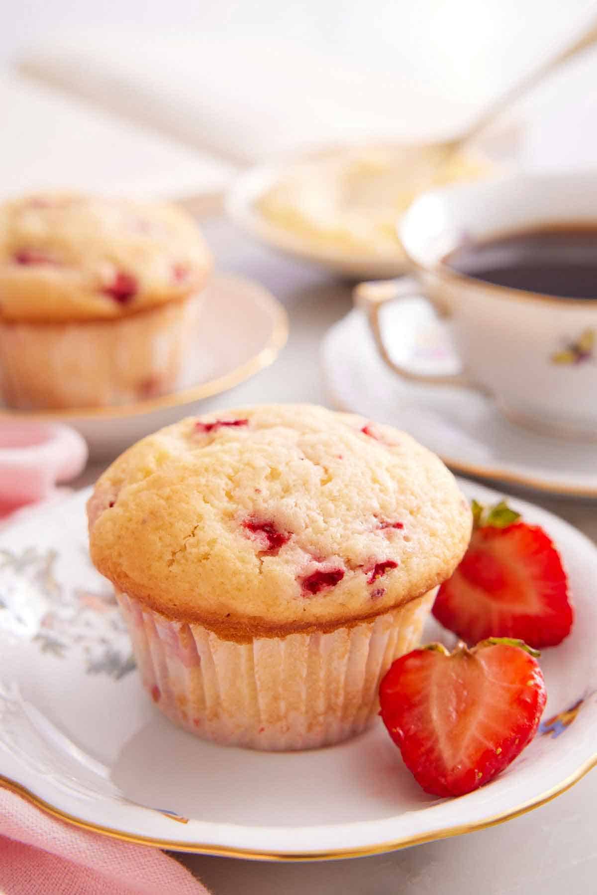 A plate with a strawberry muffin with a cut strawberry with a glass of coffee and second muffin in the background.