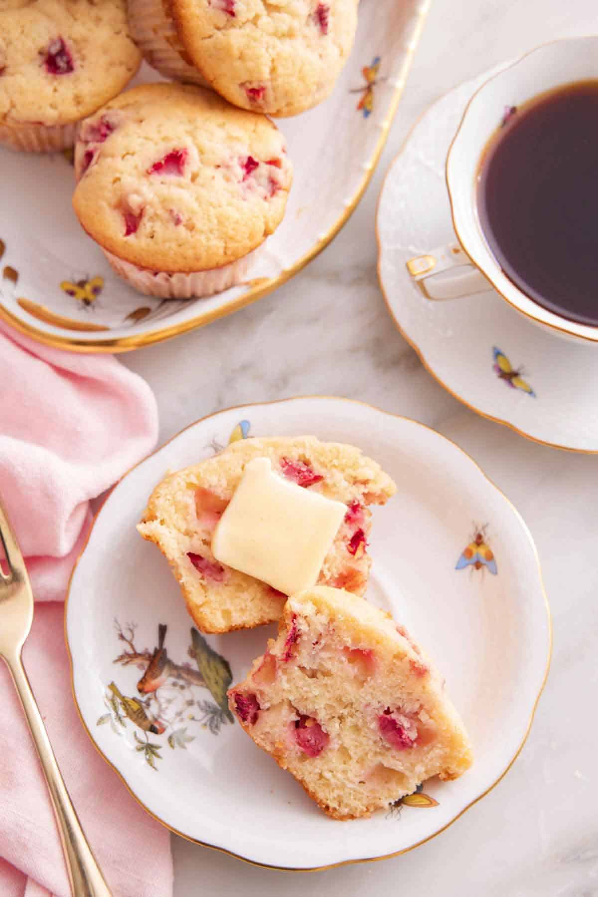 Overhead view of a plate with a strawberry muffin cut in half with a knob of butter. A cup of coffee and more muffins beside it.