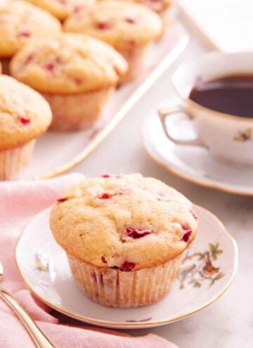 A strawberry muffin on a small plate with a cup of coffee and more muffins on a platter in the background.