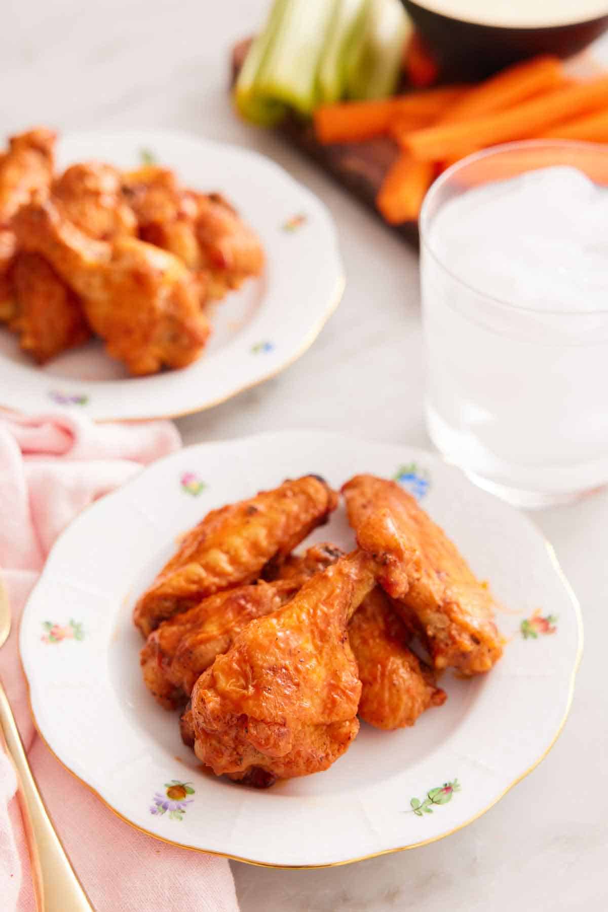 A plate with a couple of air fryer chicken wings with a glass of ice water, a plate of more wings, and a board with celery and carrots in the background.