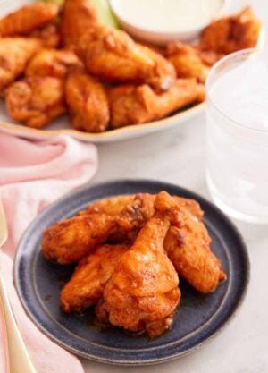A plate with multiple air fryer chicken wings with a platter of more in the background with a glass of ice water.