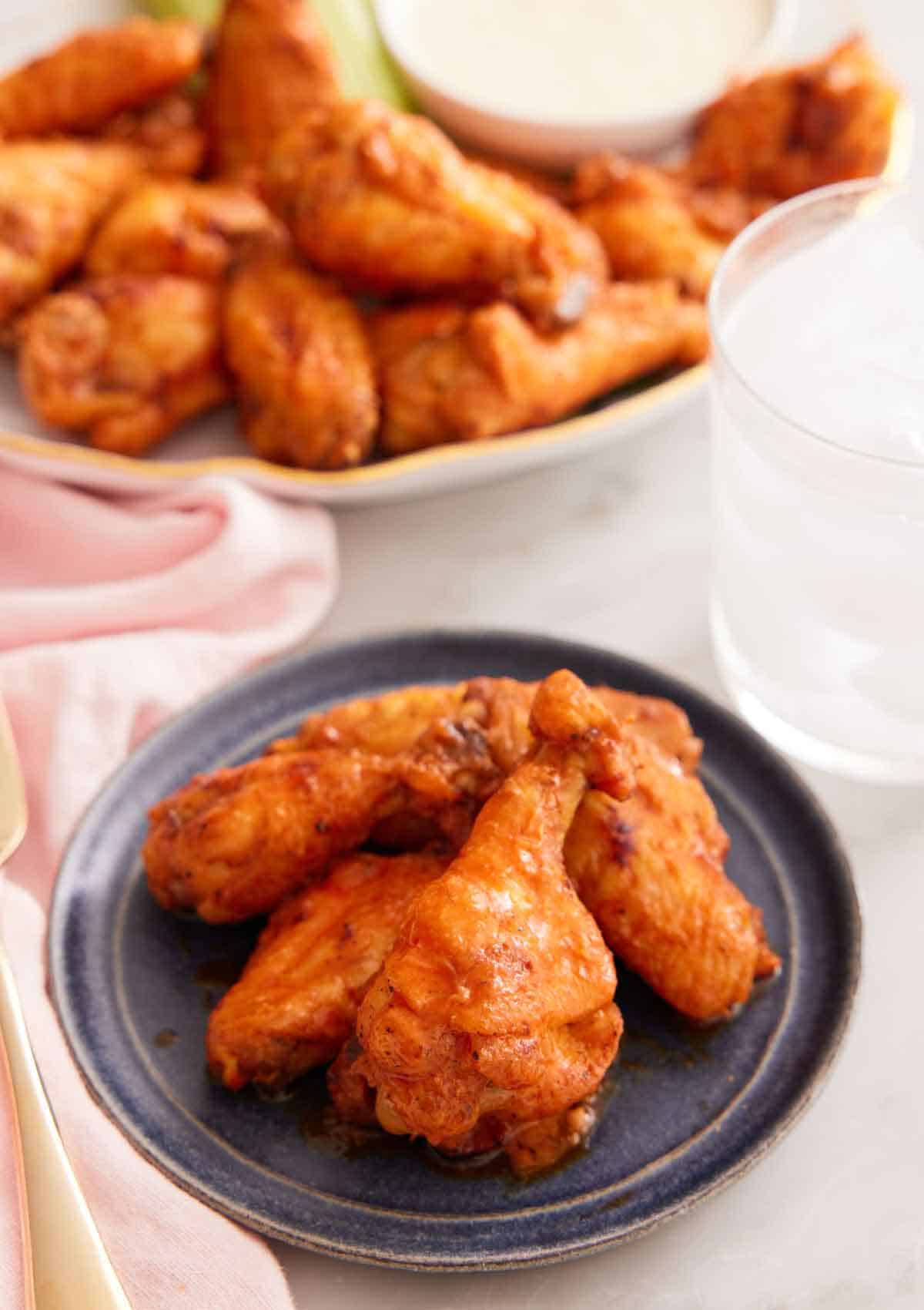 A plate with multiple air fryer chicken wings with a platter of more in the background with a glass of ice water.