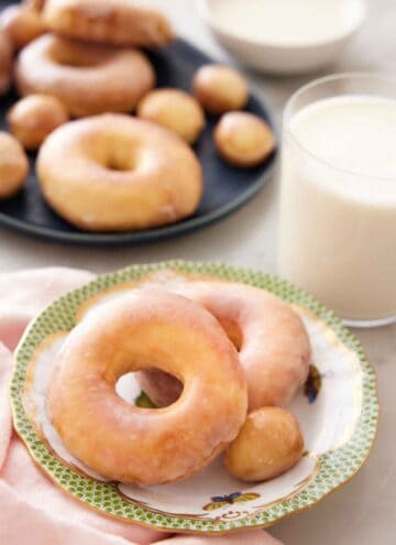 A plate with two air fryer donuts and a donut hole with a glass of milk behind it and a platter of more donuts.