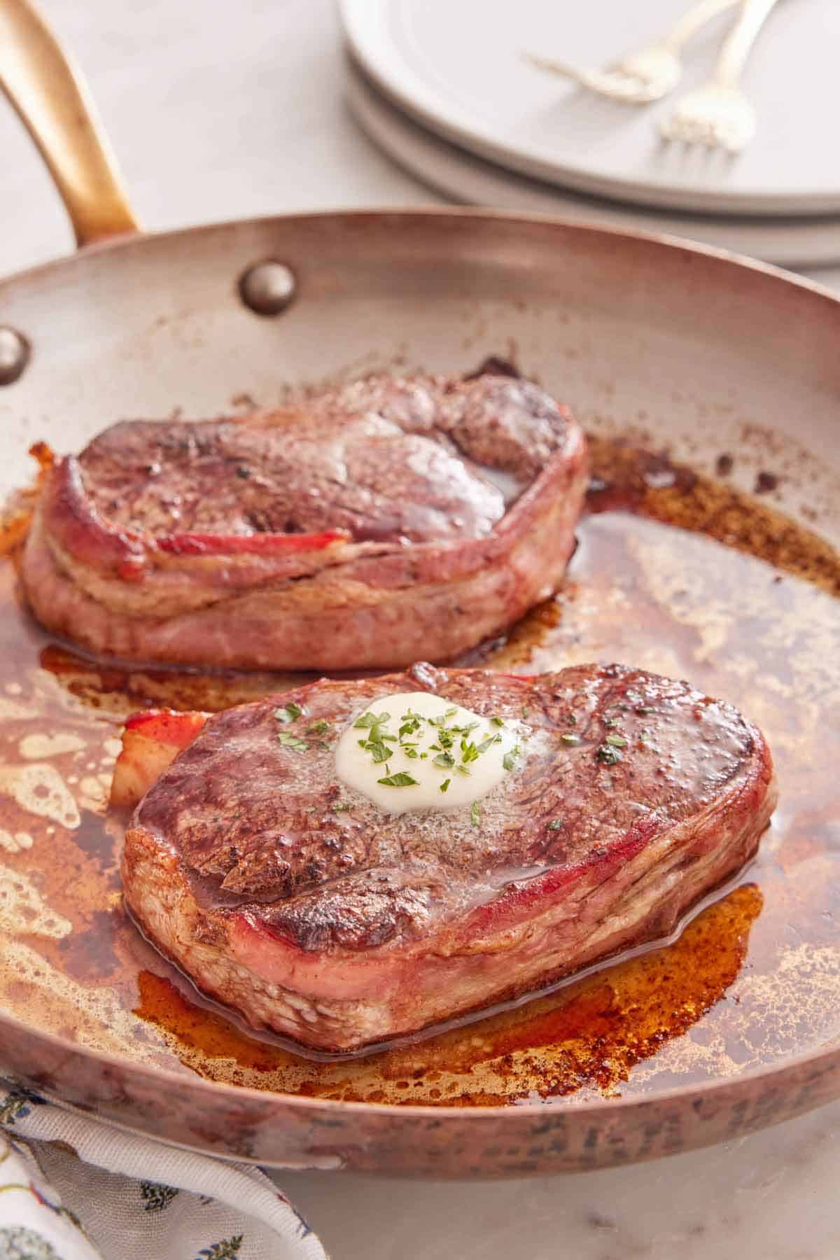 A skillet with two bacon wrapped filet mignon with one with a melted pat of butter and herbs on top.