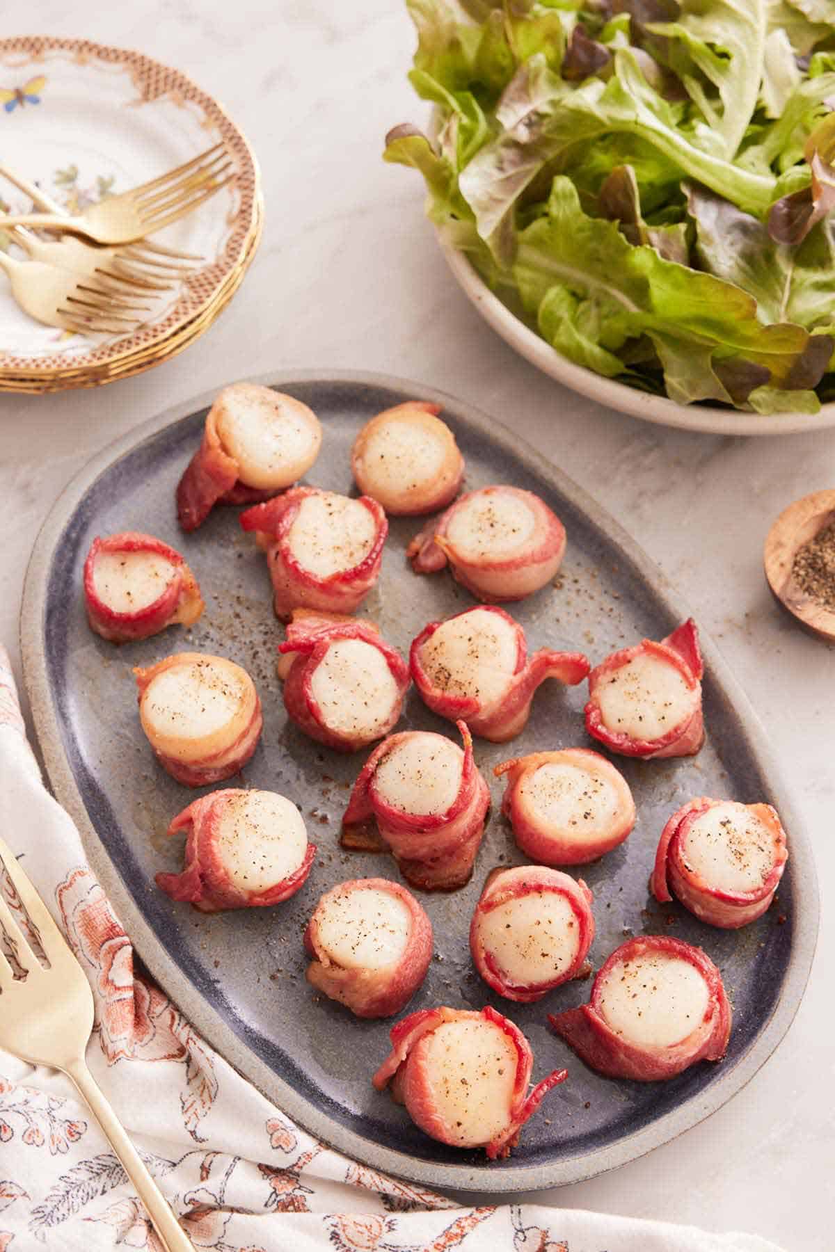 Overhead view of a platter of bacon wrapped scallops with some leafy greens on the side and a stack of plates.