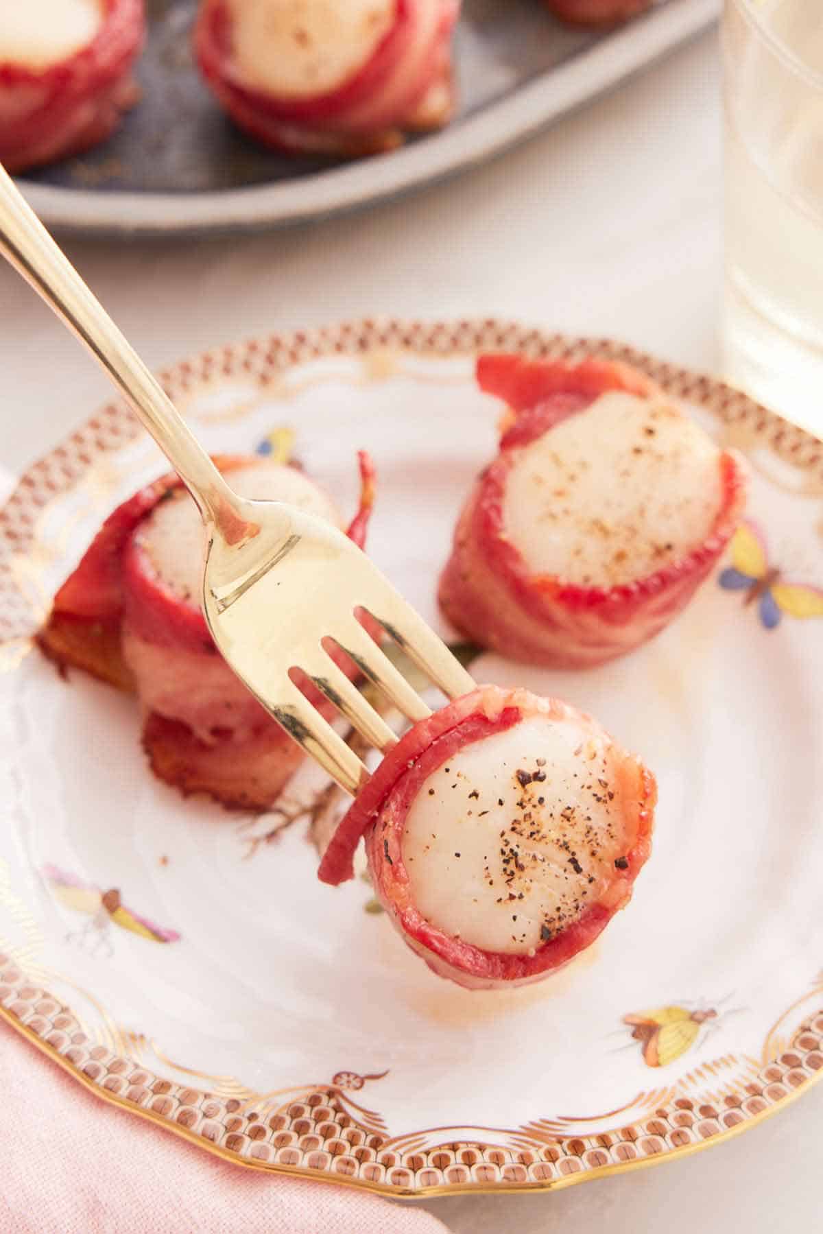 A fork lifting up a bacon wrapped scallop off a plate with two more.