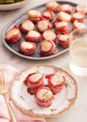 A plate with three bacon wrapped scallops with a platter in the back along with a glass of wine.