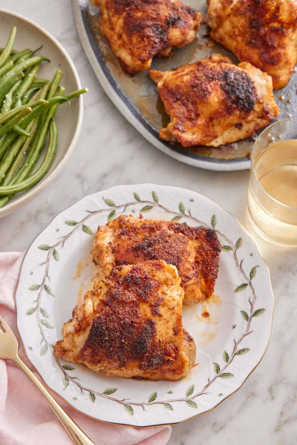Overhead view of a plate with two baked chicken thighs beside a glass of wine, platter of chicken, and green beans.