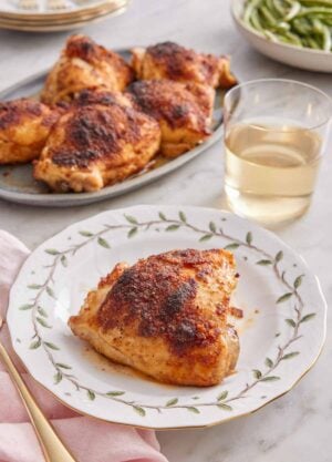 A plate with a baked chicken thigh with a glass of wine and additional chicken thighs in the background.