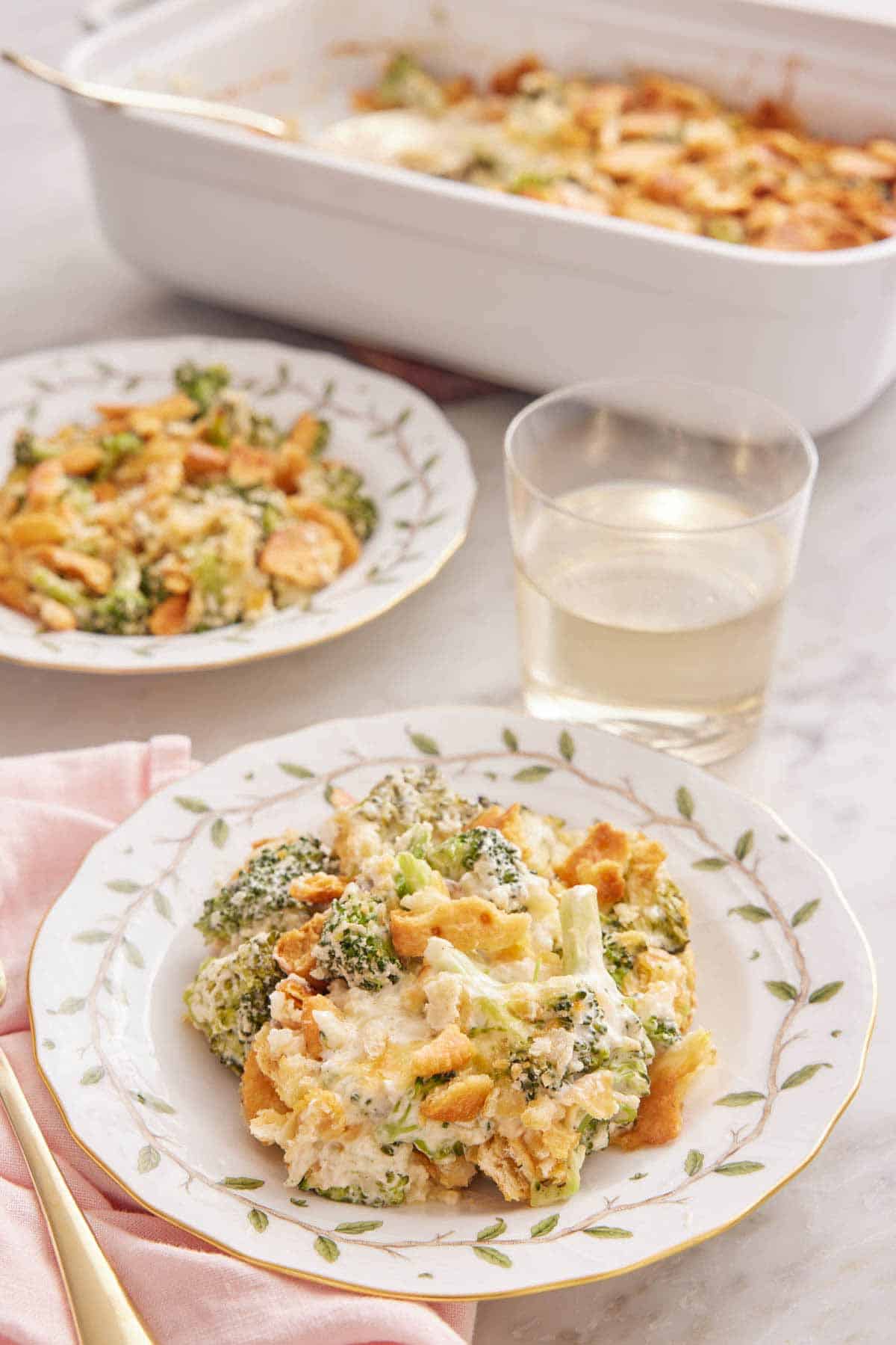 Two plates of broccoli casserole with a glass of wine inbetween and a baking dish in the back.