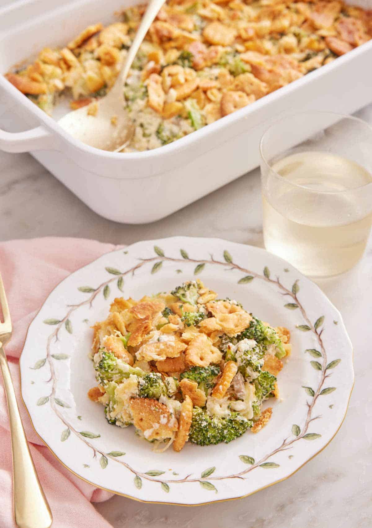 A plate with a serving of broccoli casserole with a glass of wine and baking dish in the background.