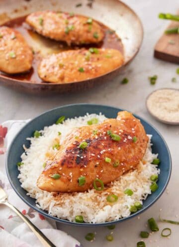 A bowl of rice with a honey garlic chicken breast on top with sesame seeds and green onions as garnish.
