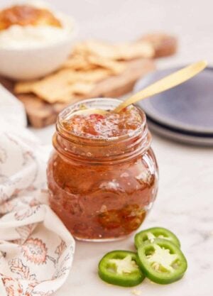 A mason jar of jalapeno jelly with a spoon inside and some sliced jalapeno in front.