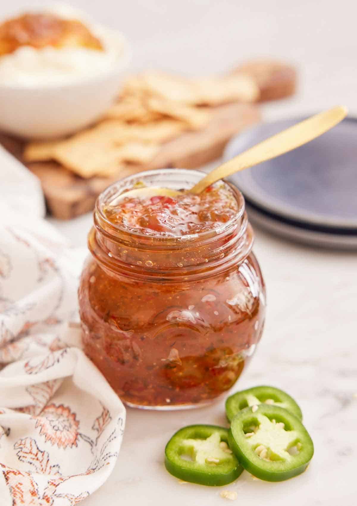 A mason jar of jalapeno jelly with a spoon inside and some sliced jalapeno in front.