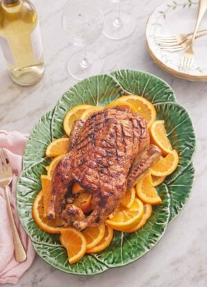 Overhead view of a green platter with a roasted duck overtop sliced oranges. Wine glasses and wine bottle in the background.