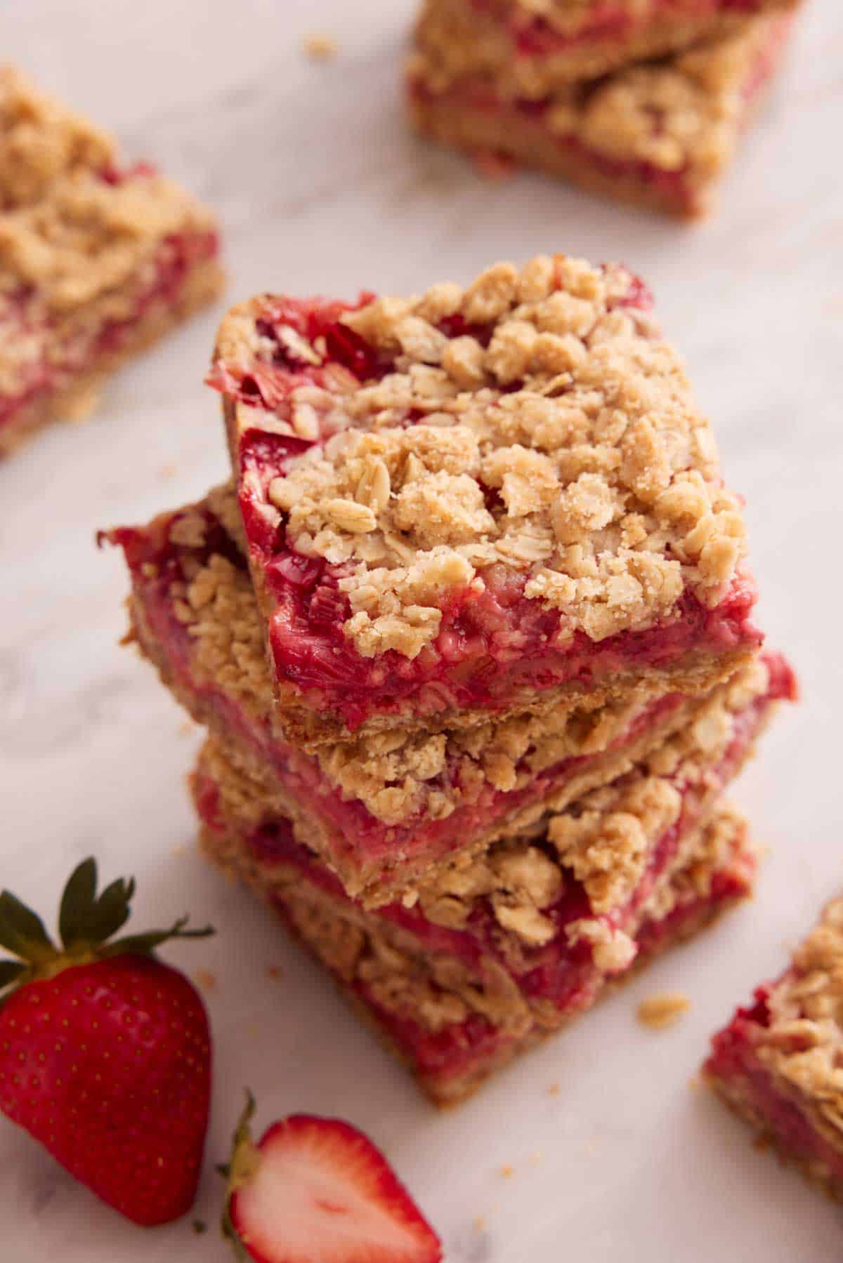 A stack of four strawberry rhubarb bars with more scattered around it and a strawberry beside it.