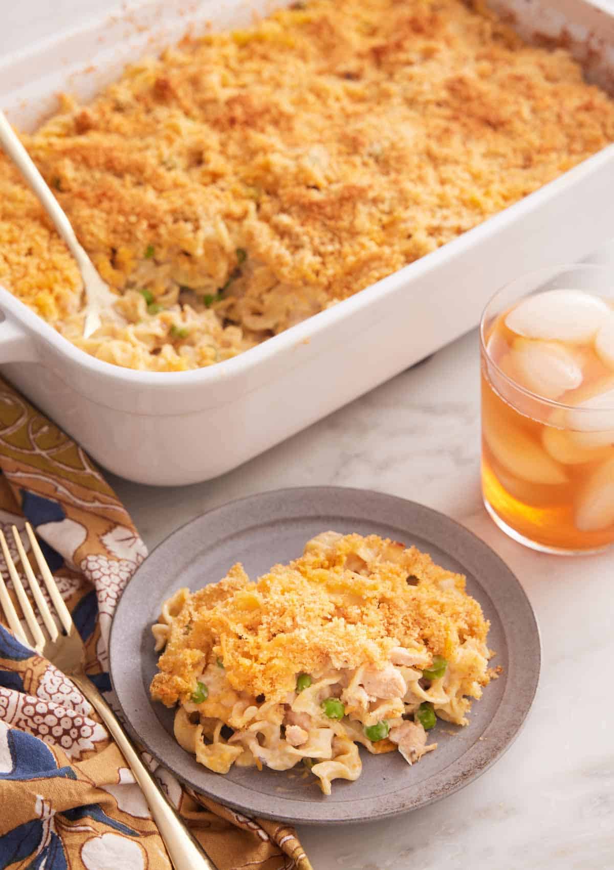 A plate with a serving of tuna casserole with a baking dish full in the back and an iced drink on the side.