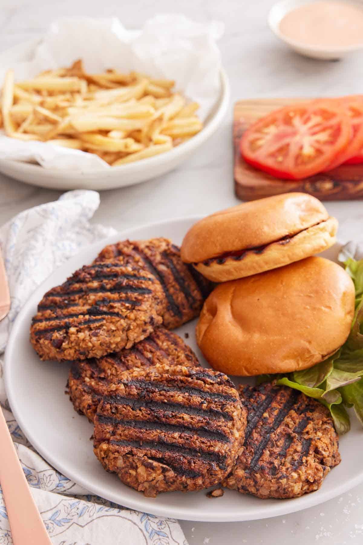 A plate with multiple veggie burger patties with two buns. Sliced tomatoes and fries in the background.