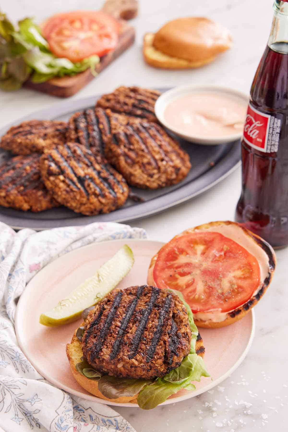 A plate with an opened veggie burger with a bottle of coke and platter of patties in the background.
