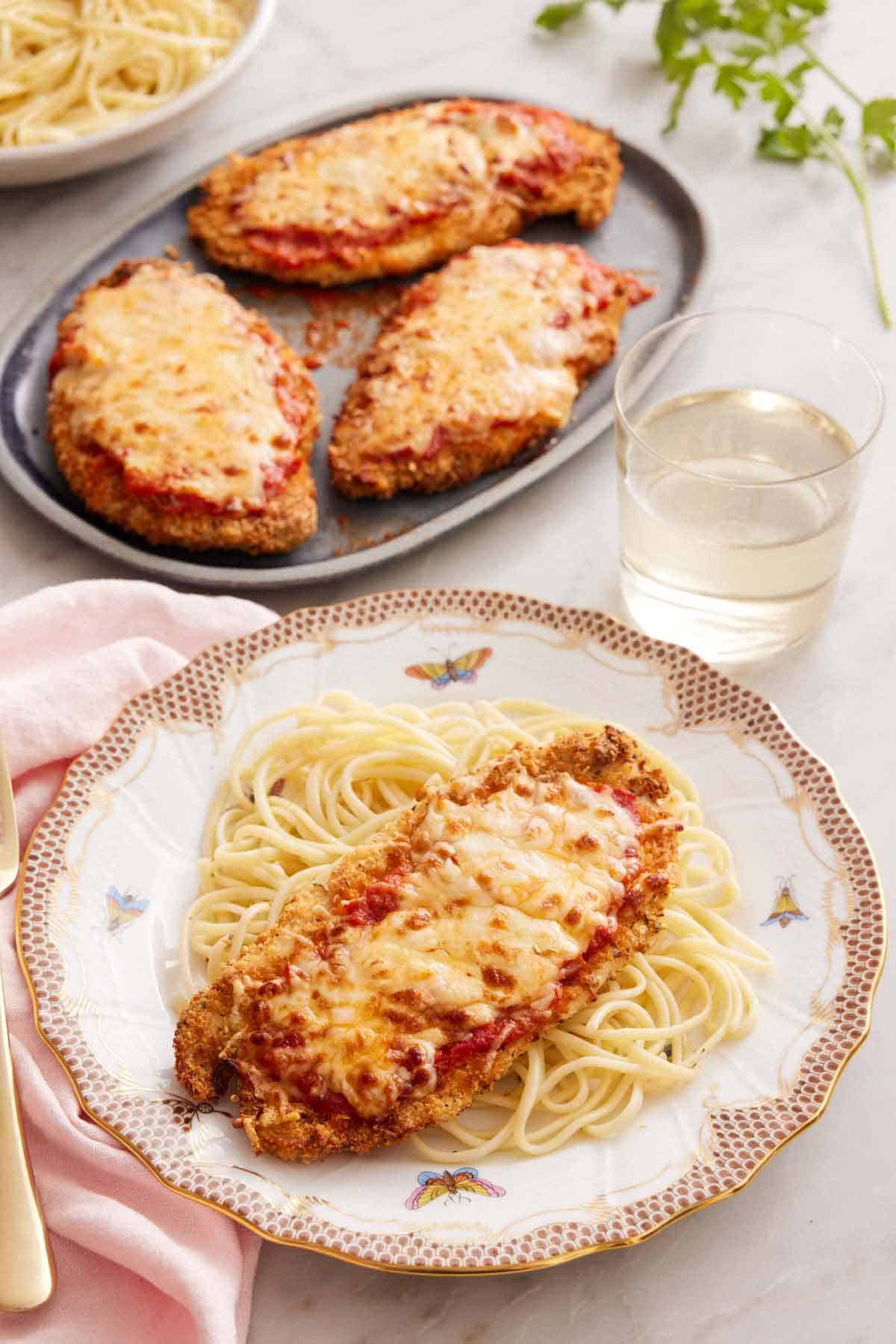 A plate with spaghetti noodles with air fryer chicken parmesan on top. A glass of wine and a platter with three air fryer chicken parmesan in the background.