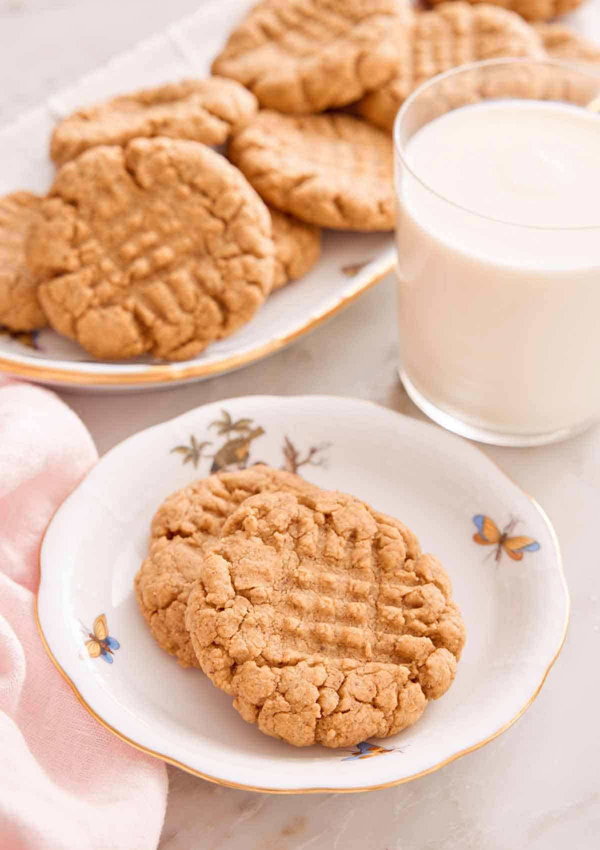 A plate with two almond butter cookies with a platter of more cookies and glass of milk behind it.