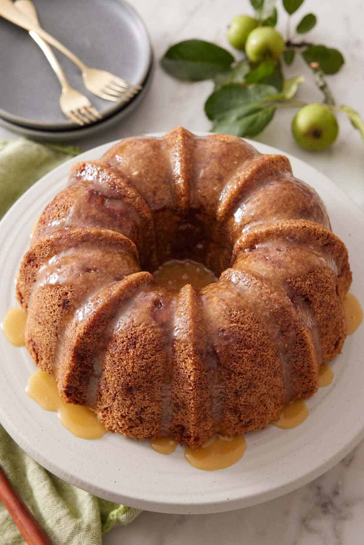 An apple bundt cake on a cake stand with a glaze drizzled over it. Apples, plates, and forks in the background.